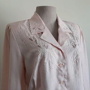 May include: A pale pink blouse with long sleeves and a collared neckline. The collar and upper chest are decorated with detailed white floral embroidery. The blouse fastens with pink buttons. The fabric has a subtle textured pattern.