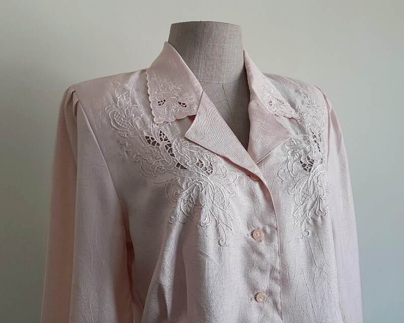 May include: A pale pink blouse with long sleeves and a collared neckline. The collar and upper chest are decorated with detailed white floral embroidery. The blouse fastens with pink buttons. The fabric has a subtle textured pattern.