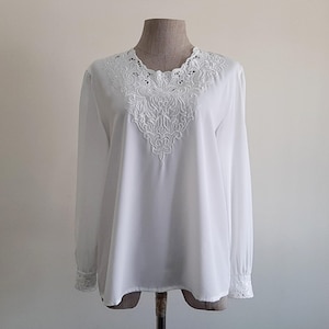 May include: A white long-sleeve blouse with intricate embroidered detailing around the neckline and down the front. The cuffs also feature delicate embroidery. The blouse is displayed on a mannequin against a neutral background.