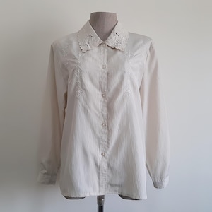 May include: Cream-colored button-down shirt with long sleeves and a decorative, openwork collar. The shirt features embroidered floral patterns on the front and cuffs. The fabric has a subtle vertical striped texture.