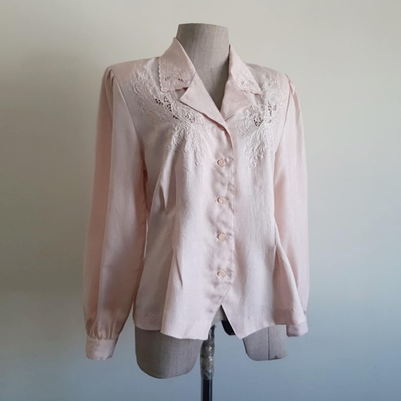 May include: A pale pink long-sleeved blouse with a collared neckline and button-down front. The collar and upper chest have delicate white embroidered detailing. The blouse is displayed on a mannequin.