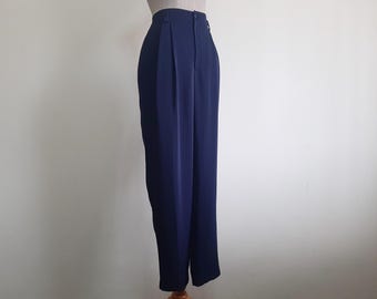 Puede incluir: Pantalones azul marino de talle alto con una cintura estructurada de un solo botón, cierre de cremallera frontal y pliegues verticales que se extienden desde la cintura por la parte delantera de cada pierna, creando una silueta entallada.
