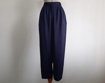 Puede incluir: Pantalones azul marino de talle alto con una cintura estructurada de un solo botón, cierre de cremallera frontal y pliegues verticales profundos que se extienden desde la cintura por las piernas, creando una silueta relajada.