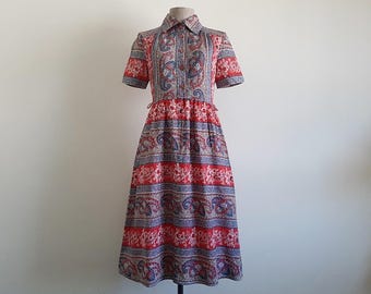 Könnte beinhalten: Ein wadenlanges Kleid mit kurzen Ärmeln und Hemdkragen, das ein sich wiederholendes horizontales Muster aus roten Blumenbändern und blauen Paisley-Motiven auf hellem Grund aufweist, mit durchgehender Knopfleiste und betonter Taille.