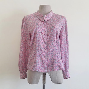 Pode incluir: Uma blusa rosa com estampa floral, gola pequena e mangas compridas. A blusa com botões apresenta um delicado padrão de pequenas flores rosa, azuis e brancas. O tecido parece leve.