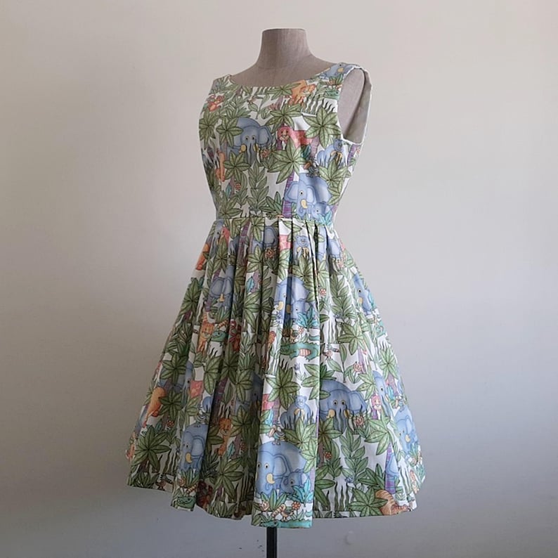 May include: A sleeveless, knee-length dress with a full skirt, displayed on a mannequin. The dress features a white background with a jungle-themed print of elephants, other animals, and lush green foliage. The dress has a fitted bodice and a flared skirt.