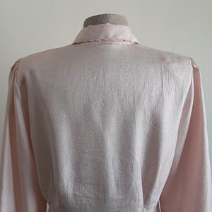 May include: A pale pink blouse with long sleeves and a collared neckline. The fabric features a delicate textured pattern. The blouse is displayed on a mannequin, showing the back and shoulder details.
