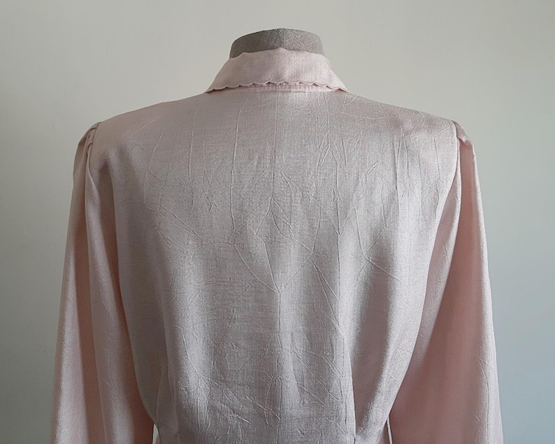 May include: A pale pink blouse with long sleeves and a collared neckline. The fabric features a delicate textured pattern. The blouse is displayed on a mannequin, showing the back and shoulder details.