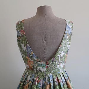 May include: A sleeveless dress with a low back, displayed on a mannequin. The fabric features a colorful jungle-themed print with animals and foliage. The dress has a pleated skirt and a scoop neckline.