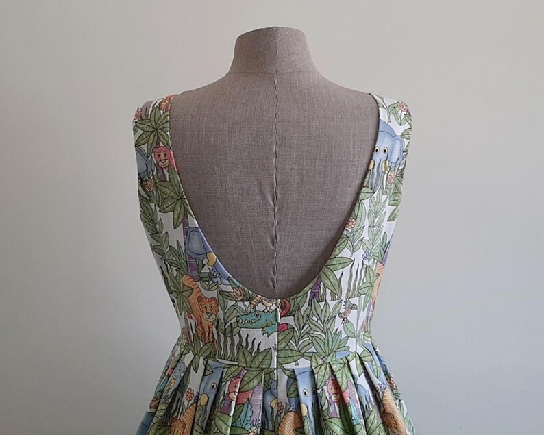 May include: A sleeveless dress with a low back, displayed on a mannequin. The fabric features a colorful jungle-themed print with animals and foliage. The dress has a pleated skirt and a scoop neckline.
