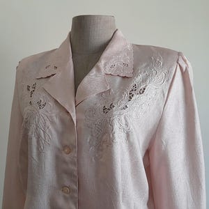 May include: Pale pink blazer with detailed floral embroidery on the lapels and upper chest. The long-sleeved jacket has a double-breasted design with two rows of buttons. The fabric has a subtle sheen, possibly satin or silk.