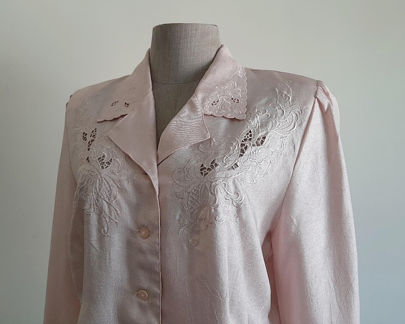 May include: Pale pink blazer with detailed floral embroidery on the lapels and upper chest. The long-sleeved jacket has a double-breasted design with two rows of buttons. The fabric has a subtle sheen, possibly satin or silk.