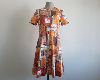 May include: A short-sleeved dress featuring a geometric block pattern in shades of orange, brown, red, and white. The garment has a round neckline, a fitted bodice, and a flared skirt that falls to the knees, displaying a repeating abstract design.