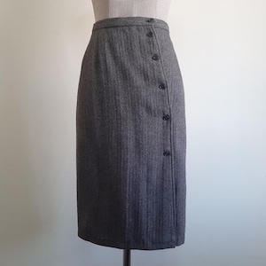 May include: A gray pencil skirt with a herringbone pattern. The skirt features a button closure on the left side and a tailored fit. The skirt is displayed on a mannequin.
