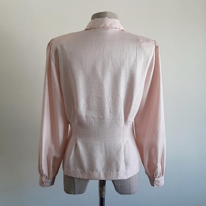 May include: A pale pink long-sleeved blouse with a collared neckline and buttoned cuffs. The back view of the blouse shows a fitted waist and a textured fabric. The blouse is displayed on a mannequin.