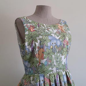 May include: A sleeveless dress featuring a jungle-themed print with elephants, lions, and monkeys among palm trees and foliage. The dress is displayed on a mannequin, showcasing its pleated skirt and scoop neckline. The fabric has a white background with colorful illustrations.