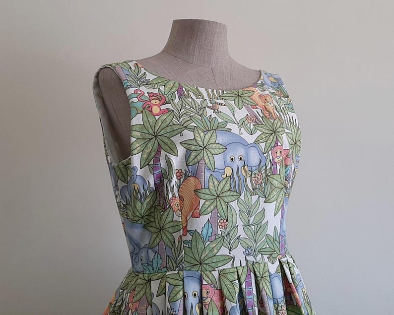 May include: A sleeveless dress featuring a jungle-themed print with elephants, lions, and monkeys among palm trees and foliage. The dress is displayed on a mannequin, showcasing its pleated skirt and scoop neckline. The fabric has a white background with colorful illustrations.