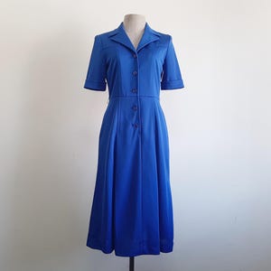 Può includere: Un abito blu brillante con maniche corte e scollo a camicia. L'abito presenta una chiusura frontale con bottoni e una gonna svasata, esposto su un manichino su sfondo bianco. L'abito è lungo fino al ginocchio.