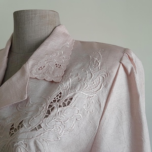 May include: Pale pink blazer with detailed floral embroidery on the collar and shoulder. The blazer has a textured fabric and a classic tailored design, displaying a vintage style. The embroidery gives a delicate, feminine feel to the garment.