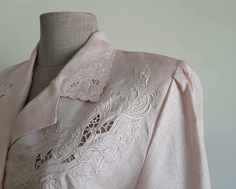 May include: Pale pink blazer with detailed floral embroidery on the collar and shoulder. The blazer has a textured fabric and a classic tailored design, displaying a vintage style. The embroidery gives a delicate, feminine feel to the garment.