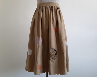 Puede incluir: Una falda midi de color tostado con cintura alta y silueta fruncida. La falda está decorada con varios parches circulares bordados en blanco, naranja y marrón, y tiene un dobladillo festoneado a lo largo del borde inferior.