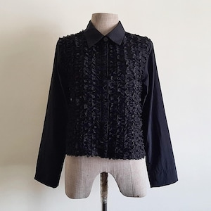 Puede incluir: Una camisa negra de manga larga con un panel frontal con volantes. La camisa tiene un cuello clásico y cierre de botones. La prenda se exhibe en un maniquí sobre un fondo neutro.