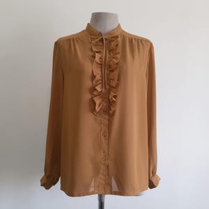 Könnte beinhalten: Langärmlige Bluse in warmem Goldbraun mit hohem Kragen und Rüschenbesatz. Die Bluse hat ein dezentes Punktmuster und geknöpfte Manschetten. Der Stoff wirkt leicht und der Stil ist vintage.