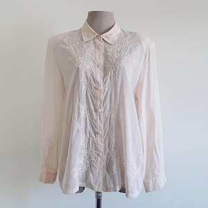 Pode incluir: Uma camisa abotoada de cor pêssego claro com mangas compridas e gola clássica. A camisa apresenta intrincados bordados florais brancos na frente e ao redor da gola. A camisa é exibida em um manequim.
