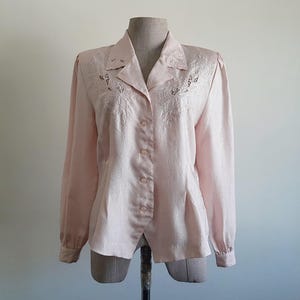 May include: A pale pink button-down blouse with long sleeves, shown on a mannequin. The blouse has embroidered detailing around the collar and upper chest. The fabric has a subtle sheen. The style is classic and feminine, suitable for various occasions.