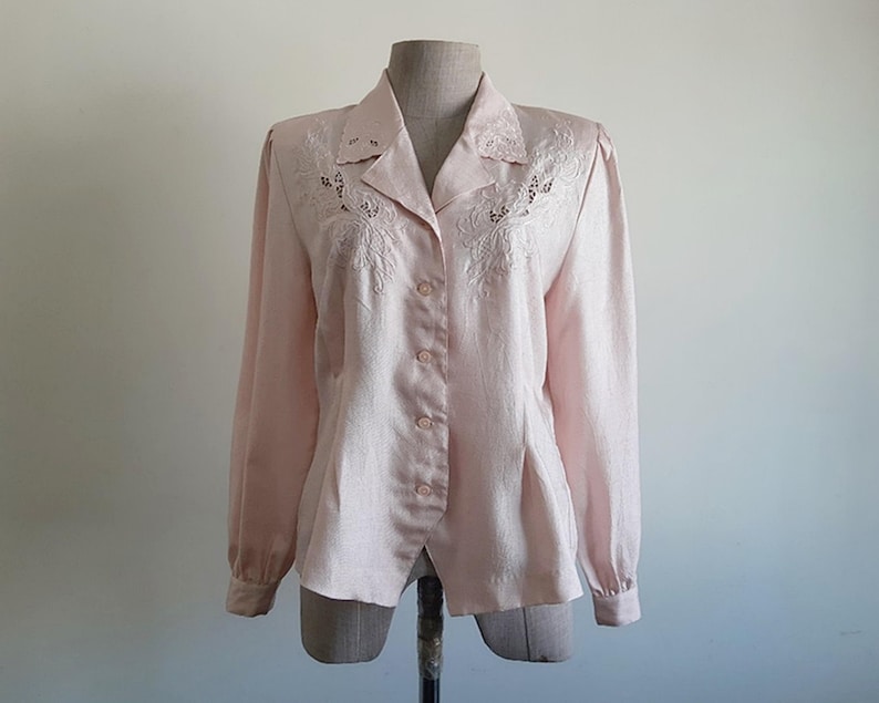 May include: A pale pink button-down blouse with long sleeves, shown on a mannequin. The blouse has embroidered detailing around the collar and upper chest. The fabric has a subtle sheen. The style is classic and feminine, suitable for various occasions.