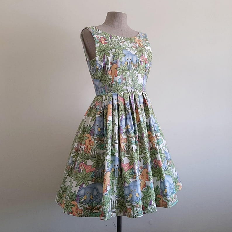 May include: A sleeveless, knee-length dress with a vibrant jungle-themed print. The fabric features elephants, lions, and other animals amidst lush green foliage. The dress has a fitted bodice and a flared skirt, displayed on a mannequin.
