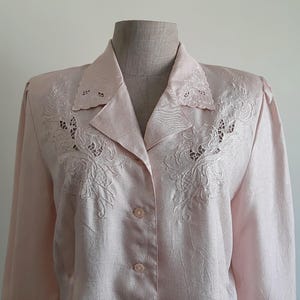May include: A pale pink button-down shirt with detailed floral embroidery on the collar and chest. The long-sleeved blouse has a classic collar and button closure. The fabric has a subtle sheen, enhancing its elegant appearance.