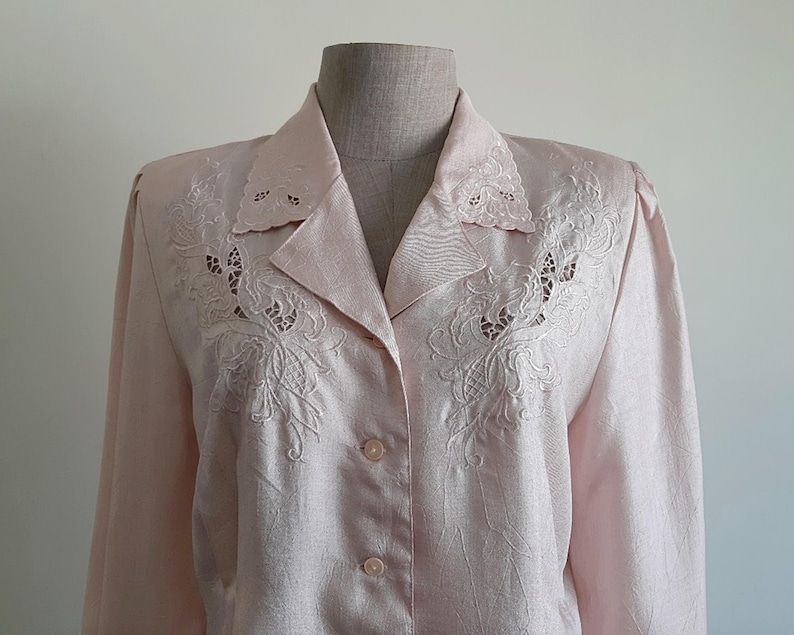 May include: A pale pink button-down shirt with detailed floral embroidery on the collar and chest. The long-sleeved blouse has a classic collar and button closure. The fabric has a subtle sheen, enhancing its elegant appearance.