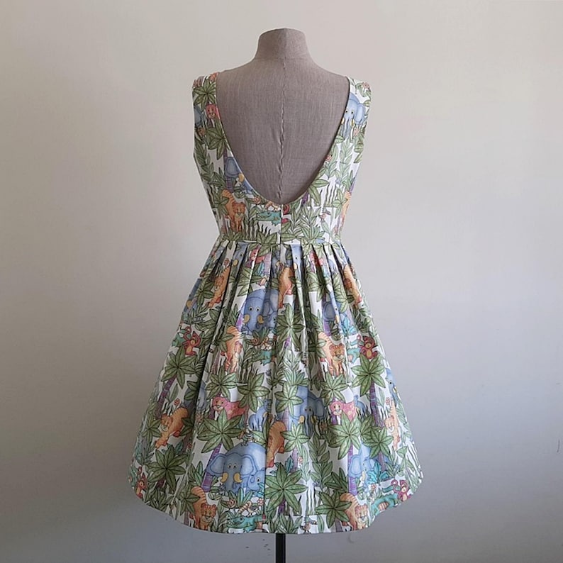 May include: A sleeveless, knee-length dress with a low back, displayed on a mannequin. The dress features a colorful jungle animal print on a white background. The print includes elephants, lions, and other animals among green foliage.