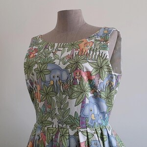 May include: A sleeveless dress featuring a jungle-themed print with elephants, lions, and other animals amidst palm trees and foliage. The dress is displayed on a mannequin, showcasing its pleated skirt and scoop neckline. The fabric has a light background.