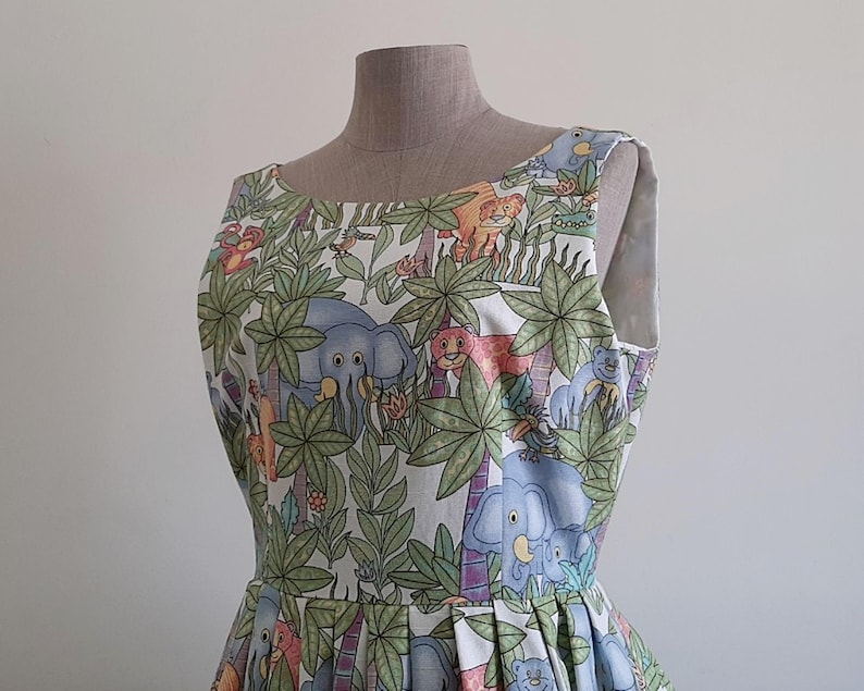 May include: A sleeveless dress featuring a jungle-themed print with elephants, lions, and other animals amidst palm trees and foliage. The dress is displayed on a mannequin, showcasing its pleated skirt and scoop neckline. The fabric has a light background.