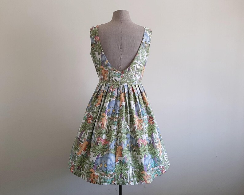 May include: A sleeveless, knee-length dress with a low back, displayed on a mannequin. The dress features a colorful jungle-themed print with elephants, giraffes, and other animals among green foliage. The skirt is pleated.