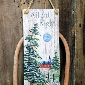 May include: A white wooden sign with the text "Silent Night" painted in black. The sign features a painting of a church with a steeple, a blue moon, and two pine trees. The sign is hanging from a twine rope.
