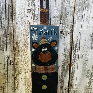 May include: A wooden sign with a black bear wearing a brown and white hat and a blue scarf. The sign says "Up North" and has snowflakes on it.