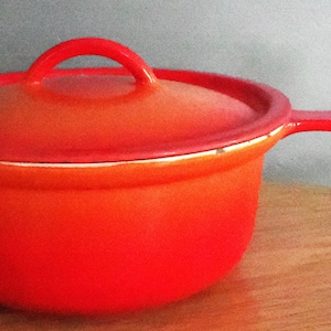 May include: A red enameled cast iron saucepan with a lid. The saucepan has a long handle and a loop handle on the lid. The pot is a vibrant red color, with a white rim. The saucepan is sitting on a wooden surface.