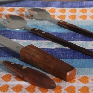 May include: A set of four salad utensils: a fork, a knife, and two spoons. The utensils have stainless steel heads and dark wood handles. The knife has a wooden sheath. The utensils are arranged on a blue and white striped cloth with orange heart patterns.