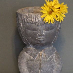 May include: A gray, weathered-looking planter shaped like a person with closed eyes and folded hands. The planter is filled with bright yellow flowers. The planter has a textured surface and is sitting on a white lace doily.