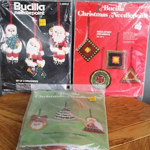 May include: Three Bucilla needlepoint Christmas ornament kits. One black package features Santa Claus designs, and a red package shows geometric shapes. A third package, green, shows painted needlepoint ornaments.