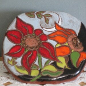 Könnte beinhalten: Eine handbemalte Keramikschale mit floralem Design. Die Schale zeigt eine große rote Blume mit braunen Details, eine orangefarbene Blume und grüne Blätter vor einem hellblauen Hintergrund. Die Schale ist oval und hat einen schwarzen Rand.
