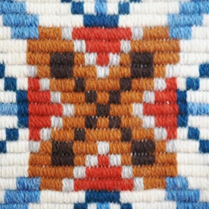 May include: A woven textile featuring a geometric pattern with a central brown block containing dark brown squares, surrounded by red, white, and blue yarn. The design consists of repeating pixelated shapes in a symmetrical arrangement of fiber.