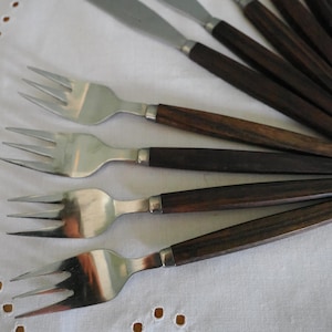 May include: A collection of vintage silverware, including forks and knives, with dark brown wooden handles and stainless steel blades. The forks have three tines. The silverware is arranged on a white tablecloth.