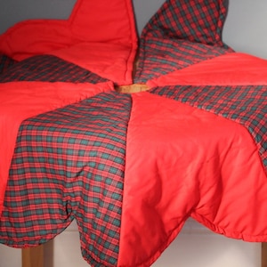 Puede incluir: Una falda de árbol de Navidad en forma de estrella, roja y a cuadros verdes. La falda presenta paneles rojos alternados y un estampado de cuadros verdes, negros y rojos. El material parece acolchado.