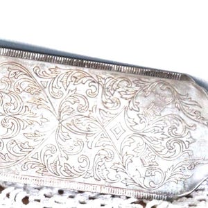 May include: A silver-toned metal cake server featuring an elongated rectangular blade with a pointed edge. The surface of the blade is decorated with intricate, engraved scrolling leaf and vine patterns across the entire flat face.