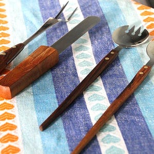 May include: A set of four stainless steel utensils featuring dark brown wooden handles secured with two metal rivets. The collection includes a two-pronged fork, a flat-edged knife, and two salad serving spoons with slotted bowls.