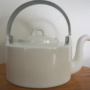 May include: A white ceramic teapot with a smooth, glossy finish, featuring a cylindrical body, a curved spout, and a matching lid with a small handle. A silver-colored metal swing handle is attached to the sides of the teapot, arching over the top.